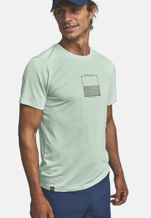 Mann trägt ein hellgrünes T-Shirt mit einer Grafik eines halb gefüllten strukturierten Glases und dem Schriftzug „HALF FULL“, kombiniert mit blauer Hose und einer Marinekappe.