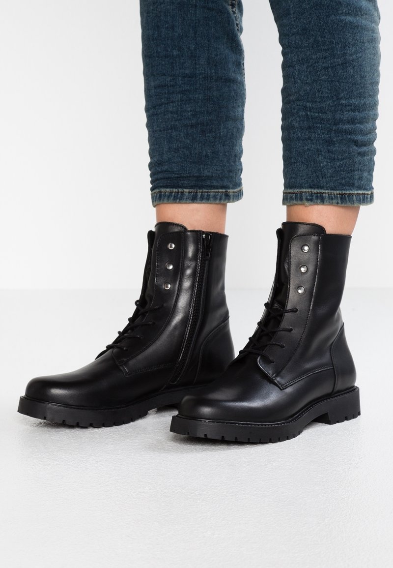 Des bottes noires en cuir jusqu'à la cheville présentent un laçage à l'avant et une fermeture éclair sur le côté, avec des œillets en argent et une semelle robuste à semelles crantées.