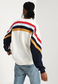 Felpa bianca con strisce orizzontali multicolori in rosso, blu, nero e senape sulle spalle e sulle maniche. Design casual, polsini a coste.