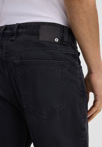Schwarze Denim-Jeans mit einem hinteren Leder-Patch, fünf Taschen-Design, sichtbaren Nähten und einer leicht verwaschenen Textur, seitlich gezeigt.
