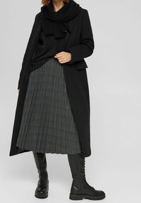 Abrigo de lana negro sobre un suéter oscuro, combinado con una falda de cuadros plisada en gris y azul. Las botas negras de cordones hasta la rodilla completan el look.