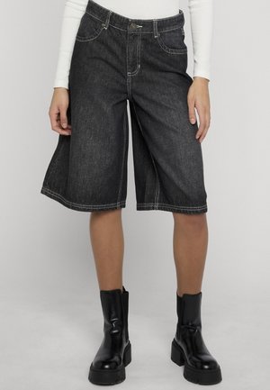 Naine kannab musti laia säärega põlvekõrguseid teksapükse valge õmblusega, mida kannab koos mustade jämedate pahkluu saabastega.