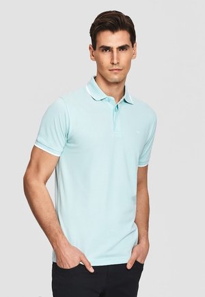 Mężczyzna w jasnoniebieskiej koszulce polo z krótkim rękawem z białym obramowaniem, stoi z rękami w kieszeniach na jednolitym szarym tle.