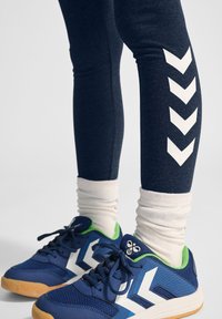 Granatowe legginsy z białym wzorem chevron po bokach, połączone z niebieskimi butami sportowymi z zielonymi akcentami i gumową podeszwą. Widoczne białe skarpety.