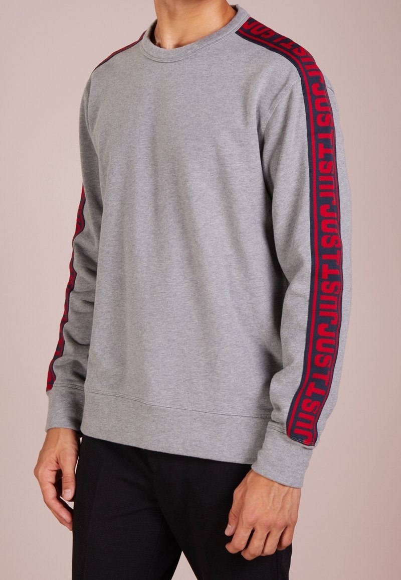 Sweatshirt gris en tissu doux, avec lettrage rouge "JUST" le long des manches. Encolure ronde classique et poignets côtelés.