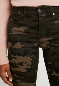 Jean imprimé camouflage en tons vert foncé et marron avec une coupe slim, doté de poches avant et d'une fermeture à bouton métallique.