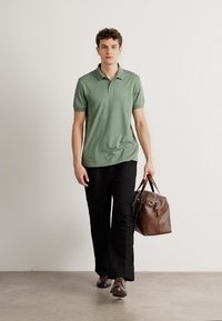 Grønn polo skjorte med krage, svarte bukser og brunt lær duffel bag. Modellen står på en jevn overflate mot en nøytral bakgrunn.