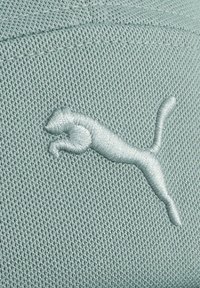 Lys teal, tekstureret stof præsenterer et broderet hvidt Puma-logo, der viser en springende kat, centralt placeret på materialet.