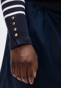 Χέρι με σκούρο δέρμα και περιποιημένα νύχια, που φοράει μπλε σκούρο πουλόβερ με λευκές ρίγες και χρυσά κουμπιά, ακουμπισμένο πάνω σε μπλε σκούρο πλισέ ύφασμα.