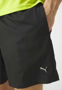 Muž v čiernych šortkách Puma a neónovo zelenom tričku, s pravou rukou opretou v vrecku.