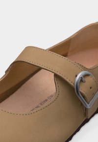 Παπούτσι Mary Jane από δέρμα ταμπά με ασημένια μεταλλική αγκράφα και εμφανείς ραφές σε λευκό φόντο.