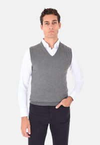 Gilet grigio lavorato a maglia con scollo a V, indossato sopra una camicia bianca con collare, abbinato a pantaloni neri. Linee pulite e morbidezza alla texture.