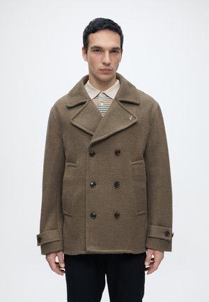 Homme portant un manteau en laine marron croisé sur une chemise rayée à col, debout devant un fond blanc uni.