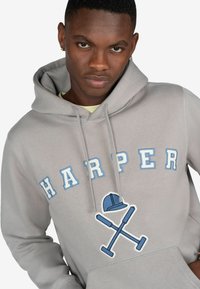 Szara bluza z kapturem z napisem "HARPER" w niebieskich literach oraz grafiką przedstawiającą niebieską czapkę i skrzyżowane narzędzia. Miękki materiał, luźny krój.