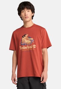 Rustfarvet bomulds t-shirt med et grafisk print af en støvle og vegetation, med "Timberland" og "BOOTS WITH ROOTS" tekst i guld nedenfor.