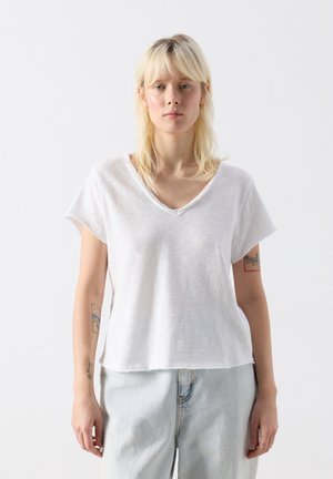 Blonde Frau, die ein locker sitzendes weißes V-Ausschnitt-T-Shirt und hellblaue Jeans trägt, vor einem schlichten weißen Hintergrund stehend.