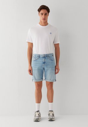 Junger Mann steht frontal mit weißem T-Shirt, hellblauen hochgekrempelten Jeansshorts, weißen Socken und grauen Turnschuhen vor einer schlichten Wand.