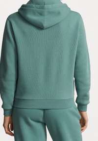 Tealfärgad sweatshirt med huva, som har ett texturerat tyg, ribbade mudd och nederkant. Baksidan är slät utan synliga sömmar eller logotyper.