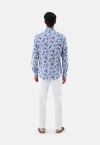 Camicia a maniche lunghe con un motivo floreale bianco e blu, abbinata a pantaloni bianchi e sandali, vista da dietro.