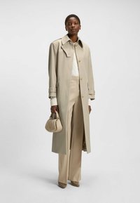 Beige Trenchcoat mit großem Kragen, Knopfverschluss und langen Ärmeln. Kombiniert mit passenden Hosen, einer cremigen Bluse und einer kleinen beigefarbenen Handtasche.