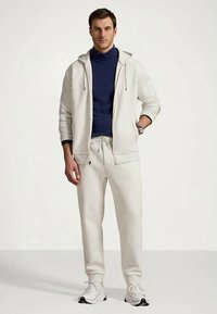 Vīrietis stāv, ģērbies beidžā kapucē, atbilstošās sporta biksēs, baltās kedās un tumši zilā turtleneck džemperī, ar vienu roku kabatā.