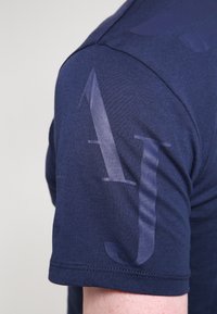 Námořnická modrá bavlněná trička s texturovaným povrchem a nadměrným, poloprůhledným designem nápisu v oblasti ramene.