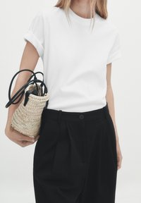 Vit bomulls T-shirt med rullade ärmar, kombinerad med svarta veckade byxor. Hand som håller en liten vävd halm väska med svarta remmar.
