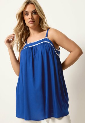 Lyshåret kvinde iført en løstsiddende blå ærmeløs kjole med hvid rickrack-trim poserende mod en lys baggrund.