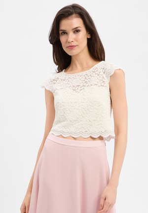 Vrouw die een witte kanten mouwloze cropped top en een lichtroze rok met hoge taille draagt, staand tegen een witte achtergrond.