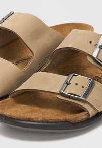 Κοντινή λήψη μπεζ σανδαλιών Birkenstock με δύο δερμάτινους ιμάντες και ρυθμιζόμενες ασημένιες μεταλλικές αγκράφες σε φελλό πάτο.