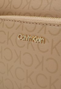 Beige kabelka s texturovaným povrchem a reliéfním logem. Zlaté kovové detaily a zipové zapínání. Viditelné značení Calvinklein.