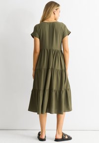 Olive green tiered dress made of soft fabric, featuring short sleeves, a round neckline, and flowing hemline. Paired with flat sandals.