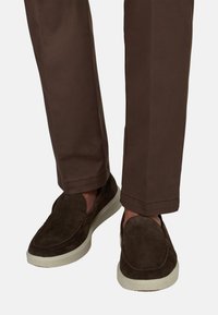 Chaussures marron en daim sans lacets avec semelles blanches, portées avec un pantalon marron à jambes droites.