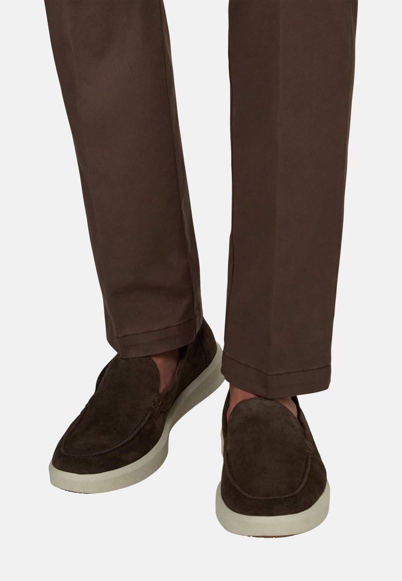 Chaussures marron en daim sans lacets avec semelles blanches, portées avec un pantalon marron à jambes droites.
