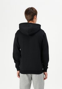 Felpa nera con cappuccio regolabile con coulisse, polsini e orlo a coste, e una texture liscia, indossata con pantaloni sportivi grigi. Vista posteriore.