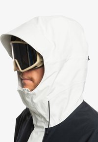 Weiße wasserdichte Kapuzenjacke mit schwarzem unteren Abschnitt und verstellbaren Zugbändern; kombiniert mit großen, getönten Skibrillen.
