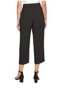 Nainen, joka on pukeutunut mustiin laajahihasiin ja mustiin korkokenkiin, seisoo selkä kameralle valkoista taustaa vasten.