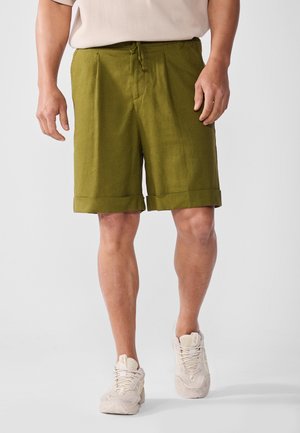 Shorts en lin verts à coupe décontractée, avec une taille à cordon de serrage, des poches latérales et des ourlets repliés. Portés avec des baskets de couleur claire.