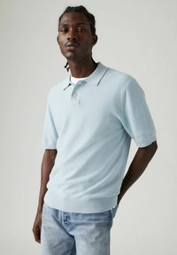 Vaaleansininen lyhytnorminen polo-paita, jossa on teksturoitu neulekuvio, taitettu kaulus ja suora helma. Yhdistetty vaaleansinisiin farkkuihin.