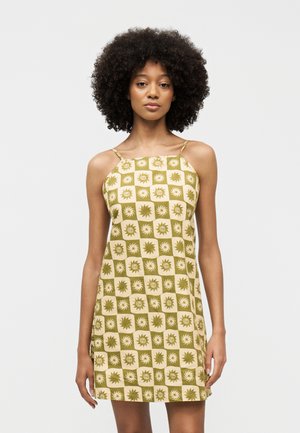 Kiharaliste juustega naine, kes kannab varrukateta beeži ja õlivihrold rohelist ruudulist kleiti päikese ja keerise mustritega.