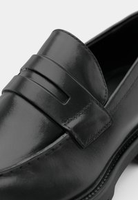 Mocassin penny en cuir noir au fini lisse, avec détails cousus et petit talon empilé, sur fond blanc.