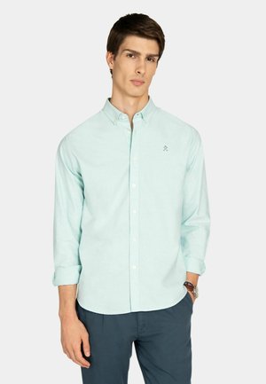 Joven con camisa azul claro de botones y mangas remangadas, y pantalones azul oscuro, de pie con una mano en el bolsillo.