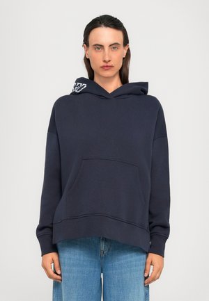 Ungdom med langt mørkt hår iført en marineblå hettegenser med lommedetaljer og lyseblå jeans, stående mot en ensfarget bakgrunn.