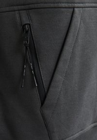 Dettaglio della tasca con zip in grigio scuro su un tessuto testurizzato, con cuciture a contrasto. Include una zip nera con linguetta.