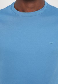 Svetlo modra majica s kratkimi rokavi, narejena iz mehkega blaga, z majhnim vezenim logotipom MK na spodnjem desnem delu. Gladka tekstura.