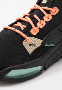 Scarpa sportiva nera con lacci arancioni, etichetta beige con logo Puma, dettagli cuciti verdi e soletta traslucida verde.