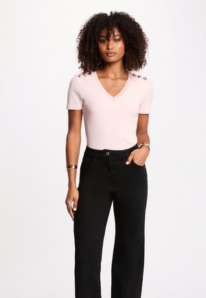 Kvinde med krøllet hår iført lyserød V-hals trøje med knapper på skuldrene og højtaljede sorte bukser, stående med den ene hånd i lommen.