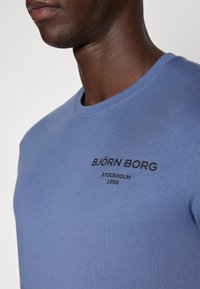 Modrá bavlněná tričko s kulatým výstřihem, krátkými rukávy a černým potiskem loga s nápisem "BJÖRN BORG STOCKHOLM 1956."