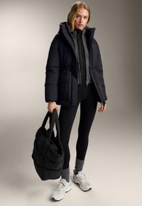 Geacă matlasată neagră cu glugă și două buzunare frontale, purtată peste un top gri și colanți negri. Poartă o geantă matlasată neagră de mână. Adidași albi.