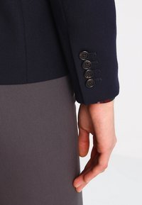 Blazer bleu marine sur mesure en tissu texturé, avec cinq boutons noirs sur la manchette et un motif intérieur visible rouge et vert.